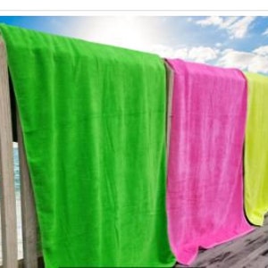 May include: A row of colorful beach towels hanging on a wooden railing. The towels are in vibrant shades of green, pink, yellow, and orange. The background shows a blue sky with clouds and the ocean. The towels are made of a soft, absorbent material.