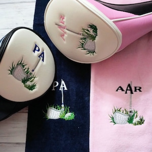 May include: Golf club head covers and matching towels. The head covers are black and pink with white accents. The towels are navy blue and pink. Each item features an embroidered golf club design and initials.
