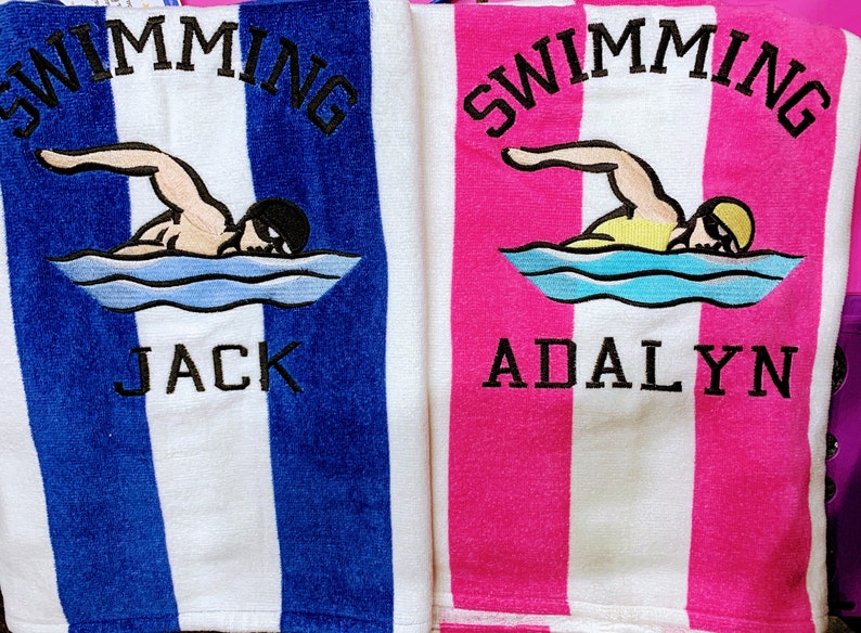 May include: Two striped beach towels, one blue and white, the other pink and white. Each towel features an embroidered swimmer graphic and the name "Jack" or "Adalyn" in black lettering, with the word "SWIMMING" above.