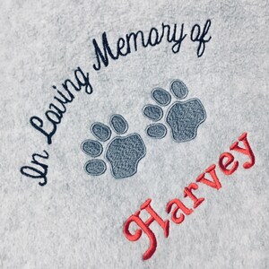May include: Light blue fabric with embroidered text and paw prints. The text reads "In Loving Memory of" in dark blue script, with two gray paw prints below. The name "Harvey" is embroidered in red.
