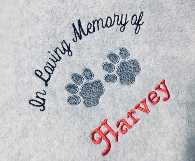 May include: Light blue fabric with embroidered text and paw prints. The text reads "In Loving Memory of" in dark blue script, with two gray paw prints below. The name "Harvey" is embroidered in red.