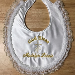 May include: White satin baby bib with gold embroidered text that reads "God Bless" above a cross and "Madelyn Lizzie". The bib is trimmed with white lace and gold accents.