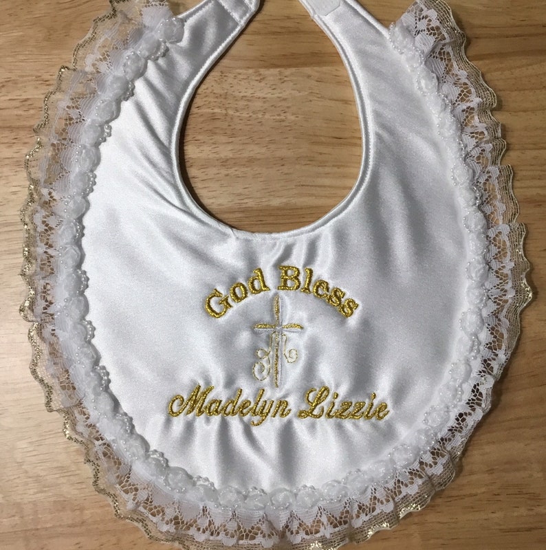 May include: White satin baby bib with gold embroidered text that reads "God Bless" above a cross and "Madelyn Lizzie". The bib is trimmed with white lace and gold accents.