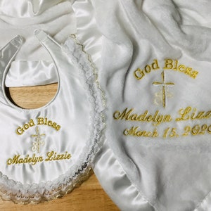 May include: White baby blanket and bib set. The blanket and bib are embroidered with gold text that reads "God Bless" and "Madelyn Lizzie March 15, 2020". The bib has a lace trim.