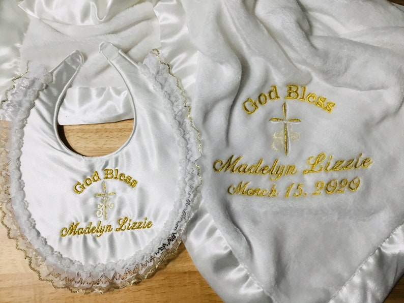 May include: White baby blanket and bib set. The blanket and bib are embroidered with gold text that reads "God Bless" and "Madelyn Lizzie March 15, 2020". The bib has a lace trim.