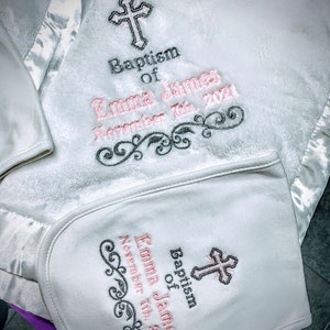 May include: White baby blanket and bib set with embroidered details. The blanket and bib feature a cross design and the text "Baptism of Emma James November 7th, 2021" in pink and black thread. The blanket has a satin trim.