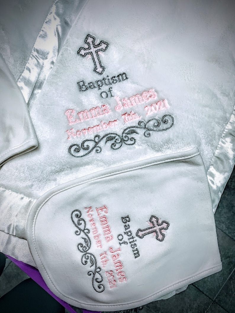May include: White baby blanket and bib set with embroidered details. The blanket and bib feature a cross design and the text "Baptism of Emma James November 7th, 2021" in pink and black thread. The blanket has a satin trim.