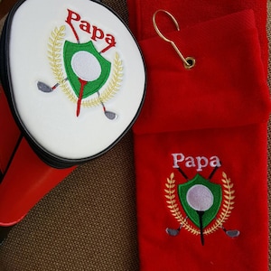 May include: A golf club head cover and a red golf towel set. The head cover is white with a red base and black zipper. Both items feature the embroidered word "Papa" above a golf-themed crest with a golf ball and clubs.