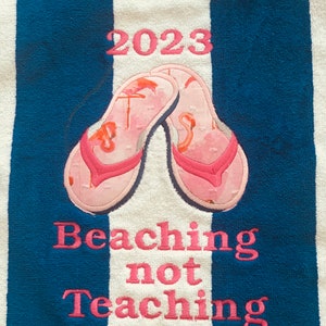 May include: Beach towel with blue and white stripes. Features pink embroidered text: "2023" above a pair of pink flip-flops with flamingo designs. Below, the text reads "Beaching not Teaching".