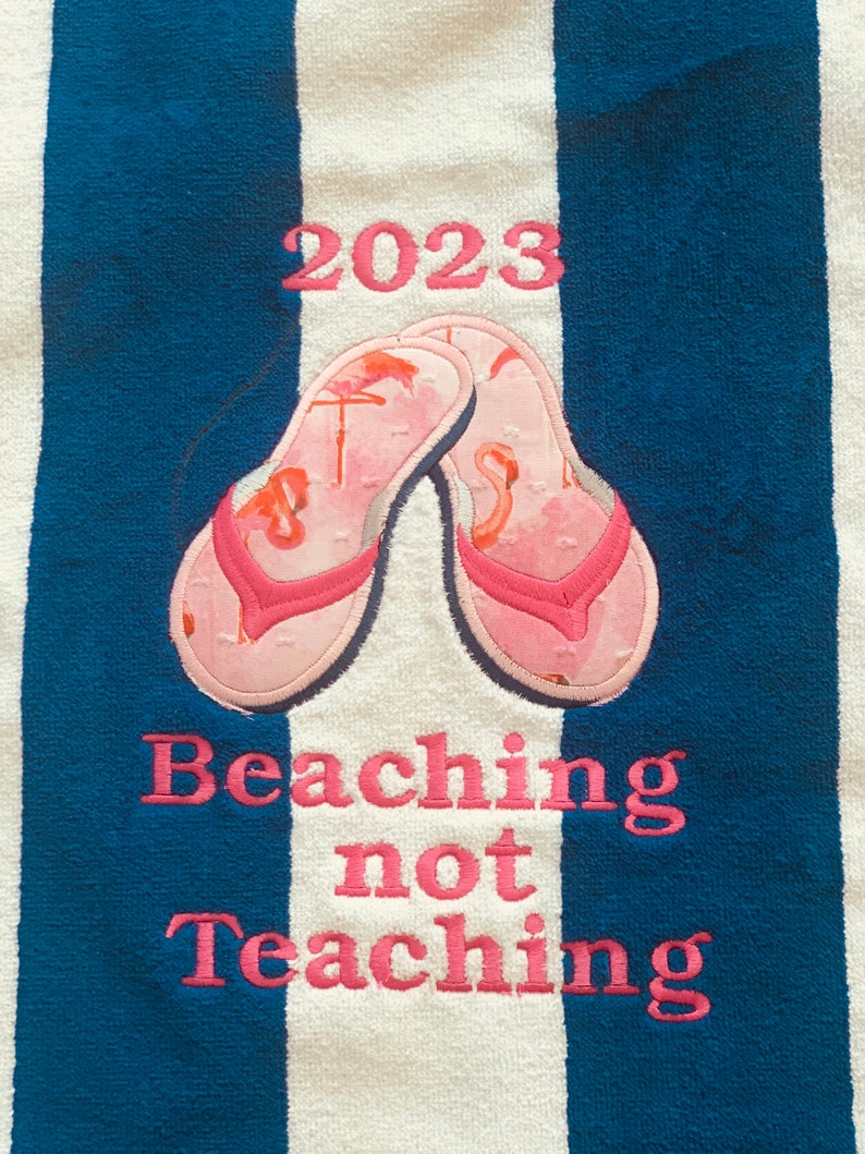 May include: Beach towel with blue and white stripes. Features pink embroidered text: "2023" above a pair of pink flip-flops with flamingo designs. Below, the text reads "Beaching not Teaching".
