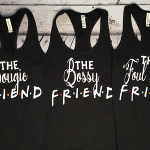 K&ouml;nnte beinhalten: Drei schwarze Tanktops mit wei&szlig;em Text. Jedes Tanktop hat einen anderen Satz: "The Bougie Friend", "The Bossy Friend" und "The Foul Mouth Friend". Das Wort "FRIEND" ist im Stil der Fernsehsendung "Friends".