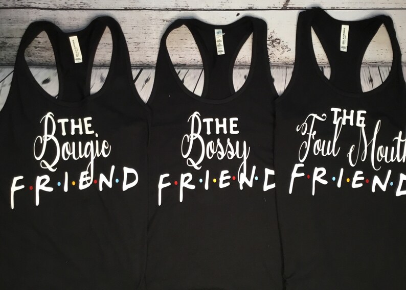 K&ouml;nnte beinhalten: Drei schwarze Tanktops mit wei&szlig;em Text. Jedes Tanktop hat einen anderen Satz: "The Bougie Friend", "The Bossy Friend" und "The Foul Mouth Friend". Das Wort "FRIEND" ist im Stil der Fernsehsendung "Friends".