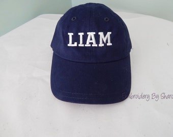 May include: Navy blue baseball cap with the name "LIAM" embroidered in white block letters. The cap has a curved brim and a classic six-panel design. The text "Embroidery By Sharon" is printed on the bottom right.
