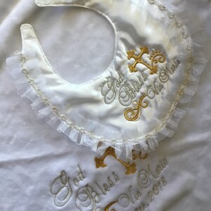 Peut inclure: Bavoir de b&eacute;b&eacute; en satin blanc avec une bordure &agrave; volants et une croix dor&eacute;e brod&eacute;e et du texte. Le bavoir indique "God Bless Mia Marie" et "April 8th, 2018". Un accessoire de bapt&ecirc;me d&eacute;licat et &eacute;l&eacute;gant.