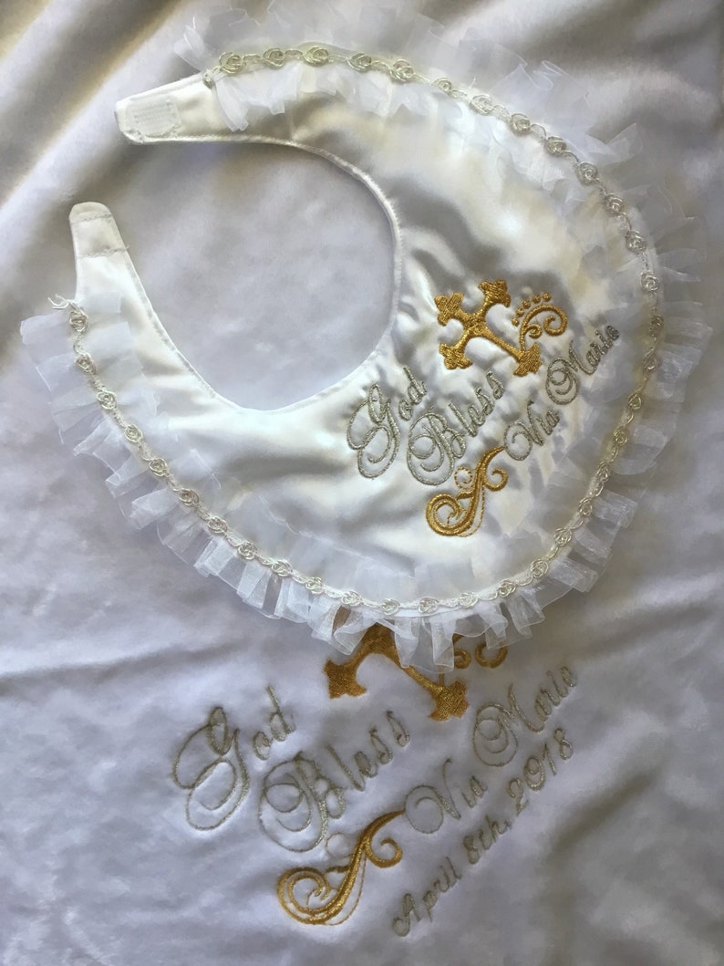 Peut inclure: Bavoir de b&eacute;b&eacute; en satin blanc avec une bordure &agrave; volants et une croix dor&eacute;e brod&eacute;e et du texte. Le bavoir indique "God Bless Mia Marie" et "April 8th, 2018". Un accessoire de bapt&ecirc;me d&eacute;licat et &eacute;l&eacute;gant.