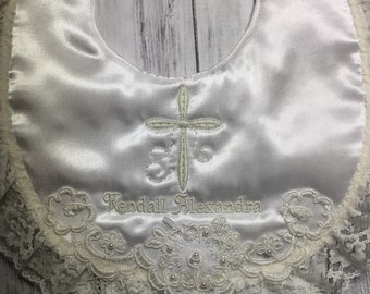May include: White satin baby bib with a cream-colored lace trim. The bib features an embroidered cross and the name "Kendall Alexandra" in a delicate script. The bottom edge is adorned with floral lace and pearl accents.