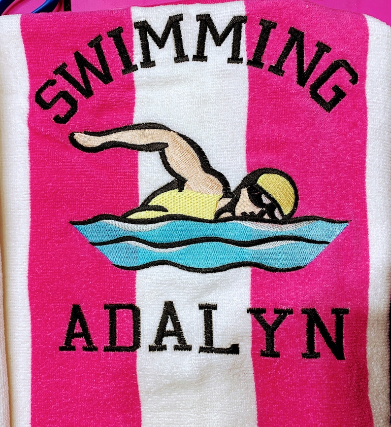 May include: A pink and white striped beach towel with the word "SWIMMING" embroidered in black at the top. Below is an embroidered swimmer graphic and the name "ADALYN" in black. The towel is made of a soft, absorbent material.