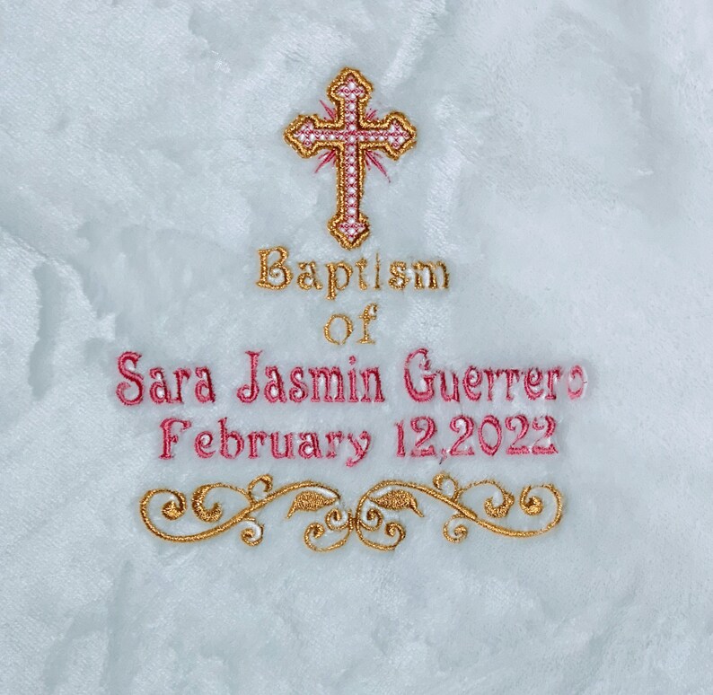 May include: Embroidered baptismal design on a white, textured fabric. The design includes a gold and pink cross, the words "Baptism of Sara Jasmin Guerrero February 12, 2022," and gold floral accents.