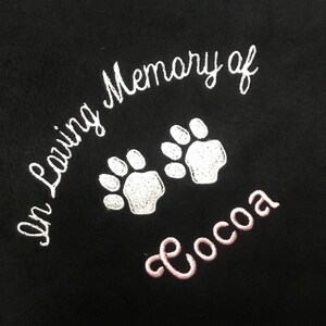 May include: Black fabric embroidered with the words "In Loving Memory of" in white script, along with two white paw prints. The name "Cocoa" is embroidered in pink script below the paw prints.