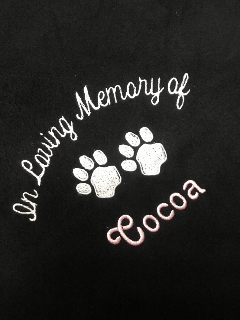 May include: Black fabric embroidered with the words "In Loving Memory of" in white script, along with two white paw prints. The name "Cocoa" is embroidered in pink script below the paw prints.