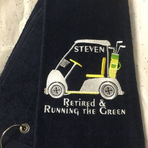 May include: Navy blue golf towel embroidered with a golf cart design. The cart is white and yellow, with the name "STEVEN" above it. The text "Retired & Running the Green" is embroidered below the cart. The towel is folded.