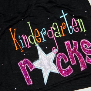 May include: Black t-shirt with the words "Kindergarten Rocks" embroidered in colorful thread and glitter. A silver star with a blue outline is centered below the text. Small, sparkly rhinestones are scattered across the shirt.
