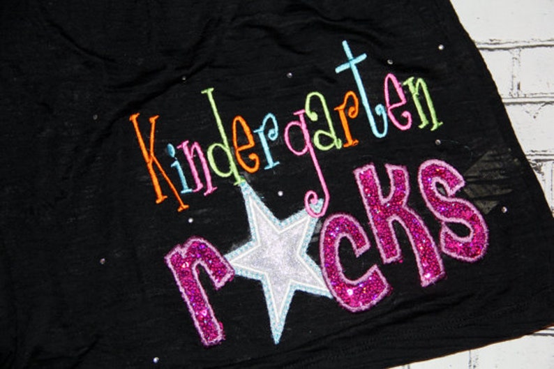 May include: Black t-shirt with the words "Kindergarten Rocks" embroidered in colorful thread and glitter. A silver star with a blue outline is centered below the text. Small, sparkly rhinestones are scattered across the shirt.