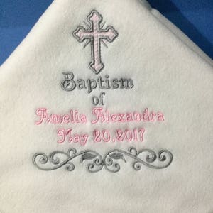 May include: White fleece baby blanket with embroidered details. Features a pink and gray cross, the words "Baptism of Amelia Alexandra May 20, 2017", and a decorative gray floral design. The blanket has a satin trim.