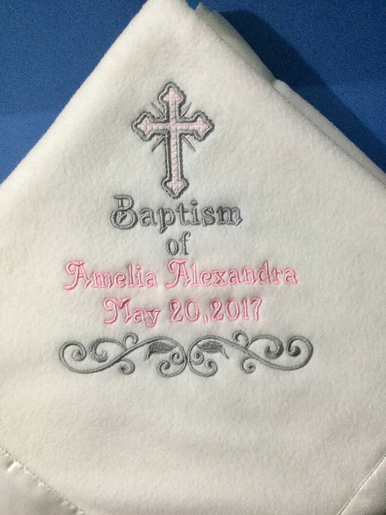 May include: White fleece baby blanket with embroidered details. Features a pink and gray cross, the words "Baptism of Amelia Alexandra May 20, 2017", and a decorative gray floral design. The blanket has a satin trim.