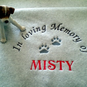 May include: A light blue fleece blanket with the embroidered text "In loving Memory of MISTY" and paw prints. A small dog figurine with brown and white markings is placed on the blanket.