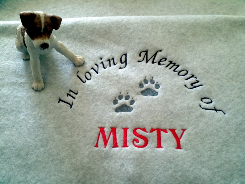 May include: A light blue fleece blanket with the embroidered text "In loving Memory of MISTY" and paw prints. A small dog figurine with brown and white markings is placed on the blanket.