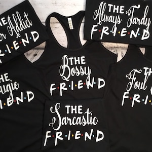 K&ouml;nnte beinhalten: Schwarze Tanktops und T-Shirts mit wei&szlig;em Text. Die Shirts tragen S&auml;tze wie "The Twitter Addict FRIEND", "The Bossy FRIEND" und "The Foul Mouth FRIEND". Das Wort "FRIEND" ist im Stil der Fernsehserie Friends gehalten.