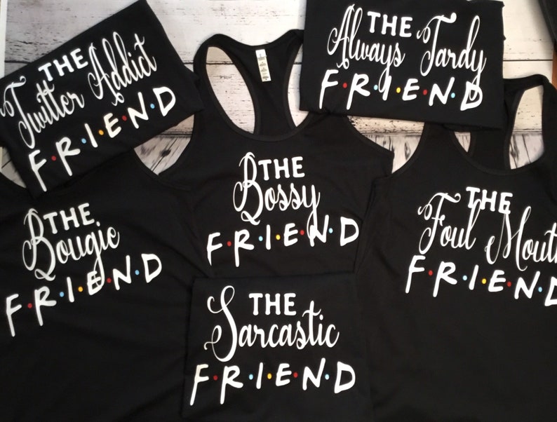 K&ouml;nnte beinhalten: Schwarze Tanktops und T-Shirts mit wei&szlig;em Text. Die Shirts tragen S&auml;tze wie "The Twitter Addict FRIEND", "The Bossy FRIEND" und "The Foul Mouth FRIEND". Das Wort "FRIEND" ist im Stil der Fernsehserie Friends gehalten.