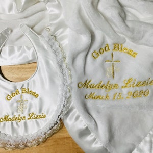 May include: A white baby blanket and bib set with gold embroidery. The blanket and bib are embroidered with the words "God Bless", a cross, the name "Madelyn Lizzie", and the date March 15, 2020. The bib has a lace trim.