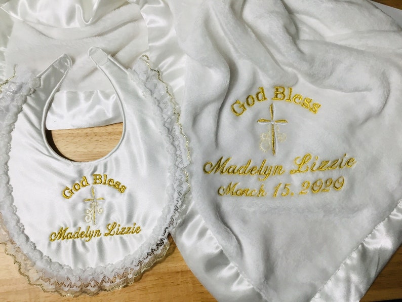 May include: A white baby blanket and bib set with gold embroidery. The blanket and bib are embroidered with the words "God Bless", a cross, the name "Madelyn Lizzie", and the date March 15, 2020. The bib has a lace trim.