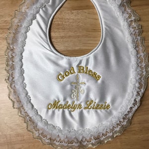 May include: White satin baby bib with gold embroidered text that reads "God Bless" and "Madelyn Lizzie" with a cross design. The bib is trimmed with white lace and gold trim.