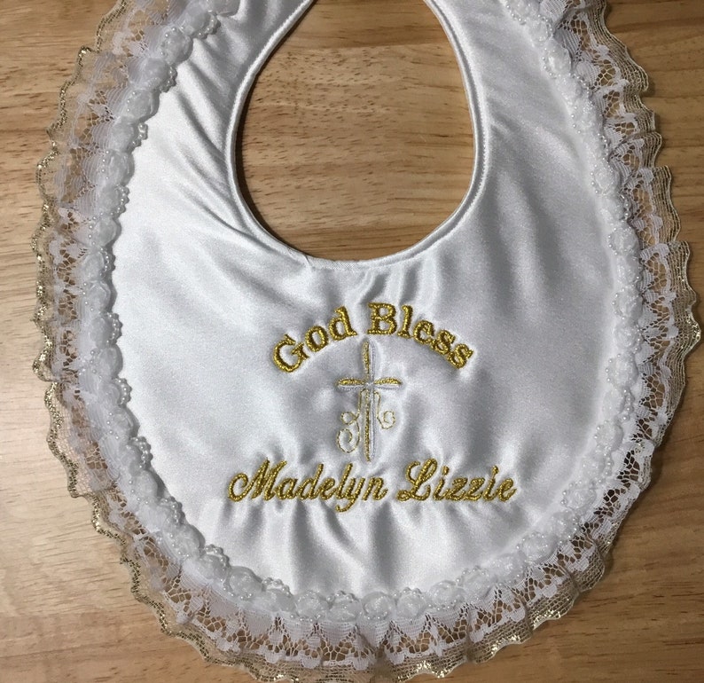 May include: White satin baby bib with gold embroidered text that reads "God Bless" and "Madelyn Lizzie" with a cross design. The bib is trimmed with white lace and gold trim.