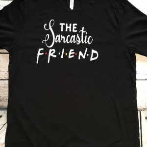 K&ouml;nnte beinhalten: Schwarzes T-Shirt mit dem Text "THE Sarcastic FRIEND" in wei&szlig;er Schrift und Blockbuchstaben. Das Wort FRIEND ist im Stil der Fernsehserie Friends gehalten, mit farbigen Buchstaben.
