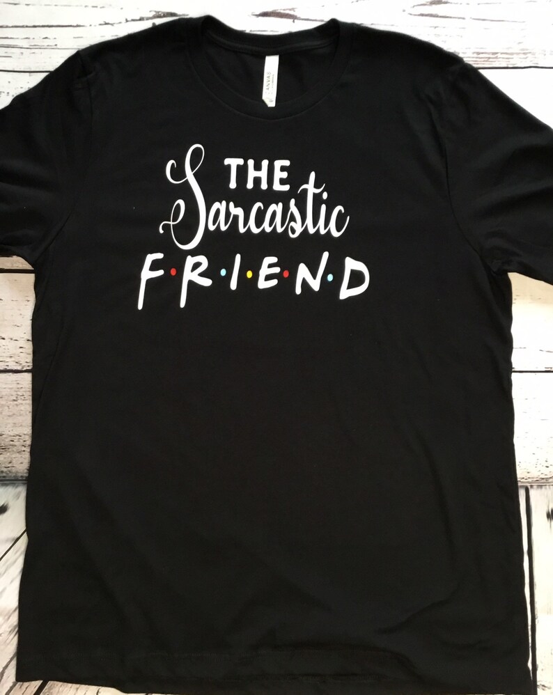 K&ouml;nnte beinhalten: Schwarzes T-Shirt mit dem Text "THE Sarcastic FRIEND" in wei&szlig;er Schrift und Blockbuchstaben. Das Wort FRIEND ist im Stil der Fernsehserie Friends gehalten, mit farbigen Buchstaben.