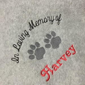 May include: A light gray towel embroidered with the words "In Loving Memory of" in black script. Two gray paw prints are embroidered below the text, and the name "Harvey" is embroidered in red script.