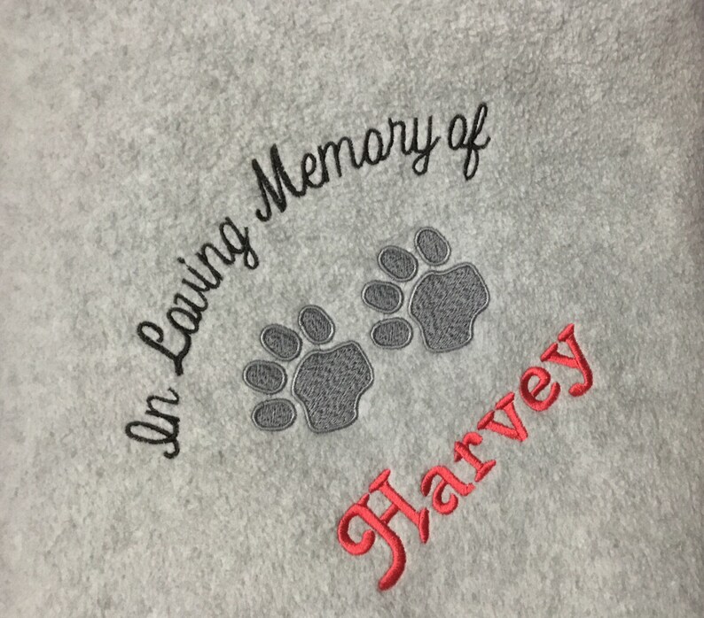 May include: A light gray towel embroidered with the words "In Loving Memory of" in black script. Two gray paw prints are embroidered below the text, and the name "Harvey" is embroidered in red script.
