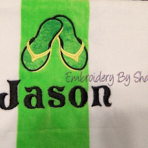 May include: White and green striped beach towel with an embroidered design of green and yellow flip-flops. The name "Jason" is embroidered in large black letters. The text "Embroidery By Sharon" is also visible.