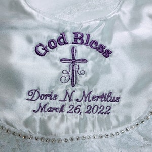 May include: White satin baptismal bib with purple embroidered text reading "God Bless", a cross, and the name "Doris N. Mertilus" with the date "March 26, 2022". The bib has a lace trim with a row of small, clear rhinestones.