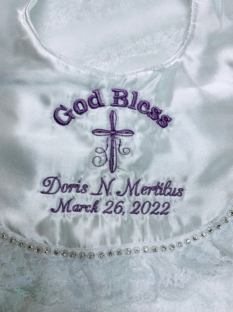 May include: White satin baptismal bib with purple embroidered text reading "God Bless", a cross, and the name "Doris N. Mertilus" with the date "March 26, 2022". The bib has a lace trim with a row of small, clear rhinestones.