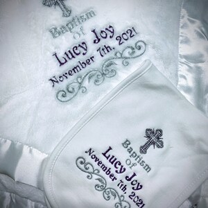 May include: White baby blanket and matching bib set. The blanket and bib feature embroidered text that reads "Baptism of Lucy Joy November 7th, 2021" and a cross design. The blanket has a satin trim.