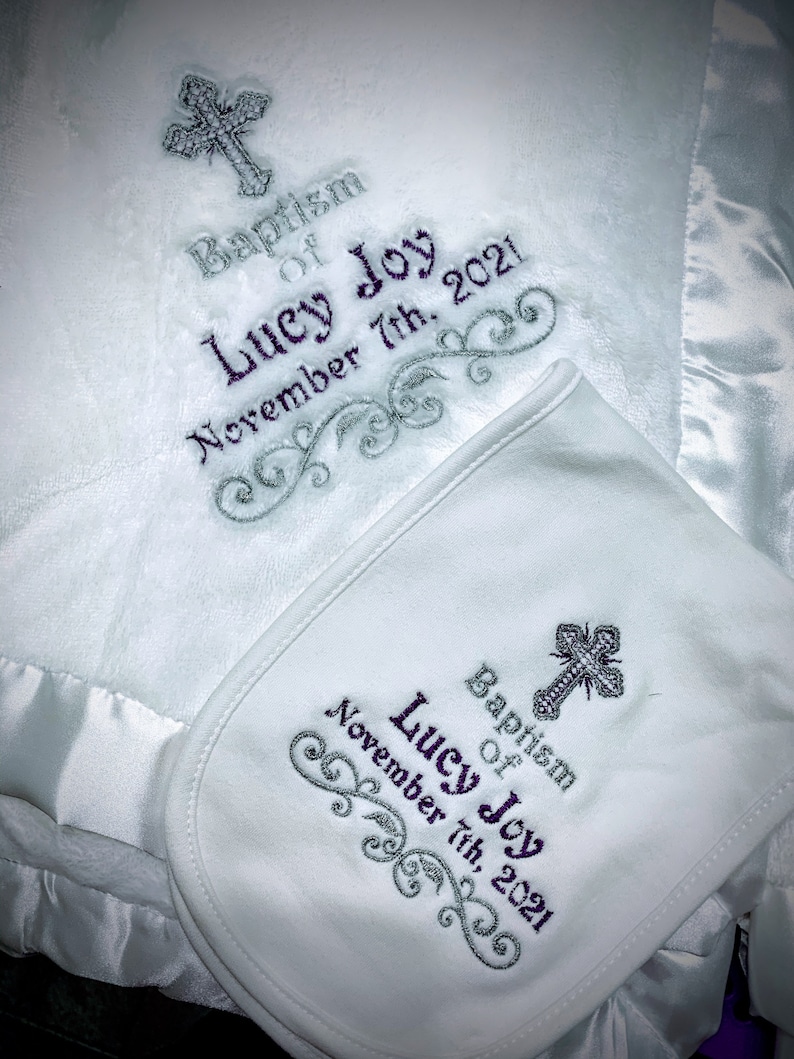 May include: White baby blanket and matching bib set. The blanket and bib feature embroidered text that reads "Baptism of Lucy Joy November 7th, 2021" and a cross design. The blanket has a satin trim.