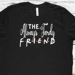 K&ouml;nnte beinhalten: Schwarzes T-Shirt mit dem wei&szlig;en Text "THE Always Tardy FRIEND". Das Wort "FRIEND" ist im Stil der Fernsehserie "Friends" gehalten, wobei jeder Buchstabe eine andere Farbe hat: rot, gelb, blau und gr&uuml;n.