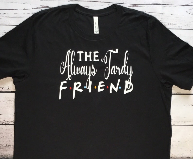 K&ouml;nnte beinhalten: Schwarzes T-Shirt mit dem wei&szlig;en Text "THE Always Tardy FRIEND". Das Wort "FRIEND" ist im Stil der Fernsehserie "Friends" gehalten, wobei jeder Buchstabe eine andere Farbe hat: rot, gelb, blau und gr&uuml;n.