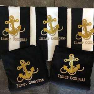 Puede incluir: Toallas de playa a rayas blancas y negras y bolsas de mano negras con un dise&ntilde;o de ancla dorada y las palabras "Inner Compass". Las toallas tienen una ancla dorada bordada. Las bolsas de mano tambi&eacute;n tienen el mismo dise&ntilde;o de ancla dorada.