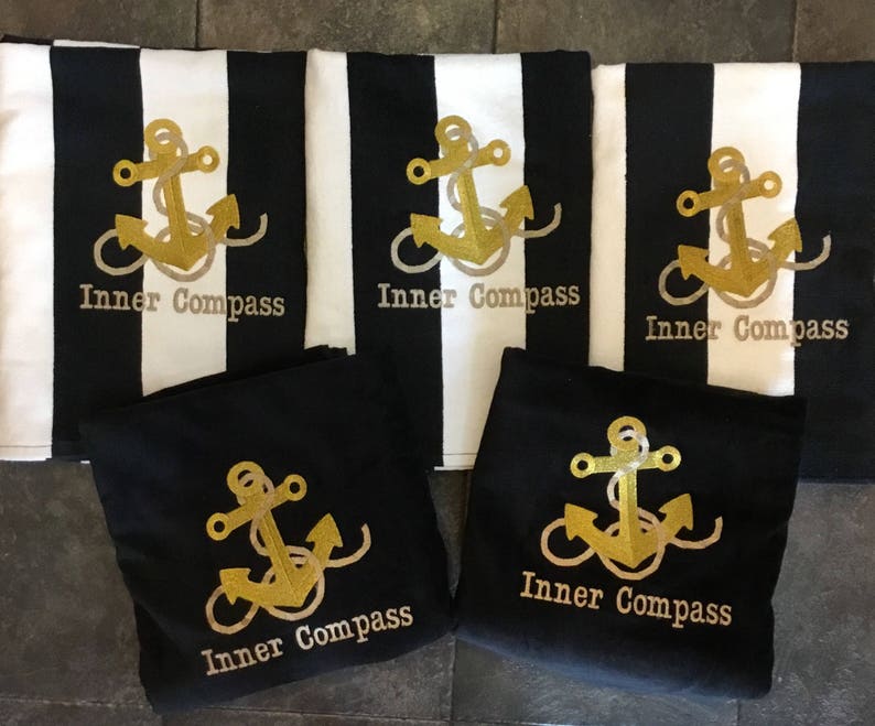 Puede incluir: Toallas de playa a rayas blancas y negras y bolsas de mano negras con un dise&ntilde;o de ancla dorada y las palabras "Inner Compass". Las toallas tienen una ancla dorada bordada. Las bolsas de mano tambi&eacute;n tienen el mismo dise&ntilde;o de ancla dorada.